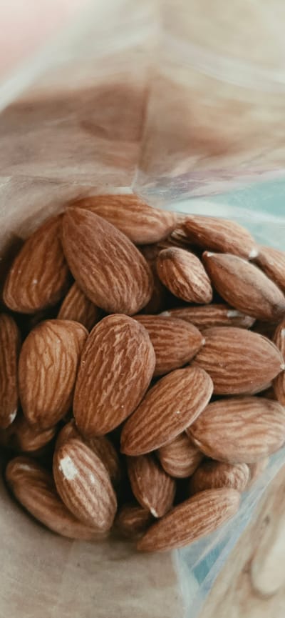 The image shows a close-up, vertical shot of a pile of almonds, likely inside a bag. The almonds are light brown with distinct ridged patterns on their shells. The focus is on the texture and shape of the nuts, with some appearing slightly lighter or darker due to lighting and natural variation. The background is blurred and appears to be the translucent plastic of the bag, with hints of muted colors like blue and beige. There is no information about Lipetsk, Russia, or any people present in the image.