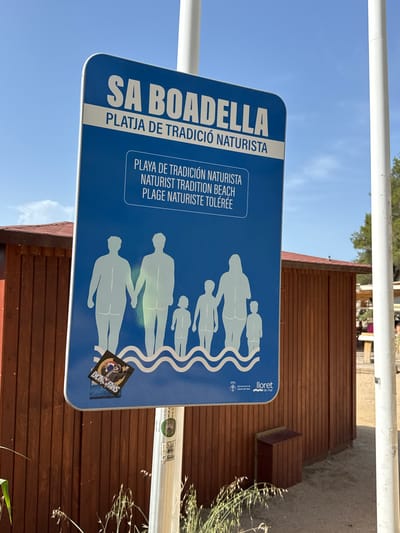 A blue and white sign is mounted on a white pole in front of a wooden building. The sign has text and an illustration of people. The text on the sign reads "SA BOADELLA PLATJA DE TRADICIÓ NATURISTA" and below that, "PLAYA DE TRADICIÓN NATURISTA NATURIST TRADITION BEACH PLAGE NATURISTE TOLÉRÉE." The illustration depicts a family group of six figures, all in silhouette, standing in water. They are holding hands. The figures are two adults, two children, and two smaller children or infants. Below the illustration is a logo for "Ajuntament de Lloret de Mar." A sticker is attached to the bottom left corner of the sign. The wooden building behind the sign has a red roof and appears to be a beach hut or changing room. The ground in front of the building is sandy. The sky is blue and clear.