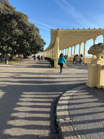 An outdoor walkway features a pergola structure with evenly spaced pillars casting long shadows on the pavement. People are walking along the walkway under the pergola. A large tree with dense foliage sits beside the walkway. The sky is clear blue with contrails. A stone curb separates the walkway from a raised platform. The scene is bathed in sunlight, creating strong contrasts between light and shadow.