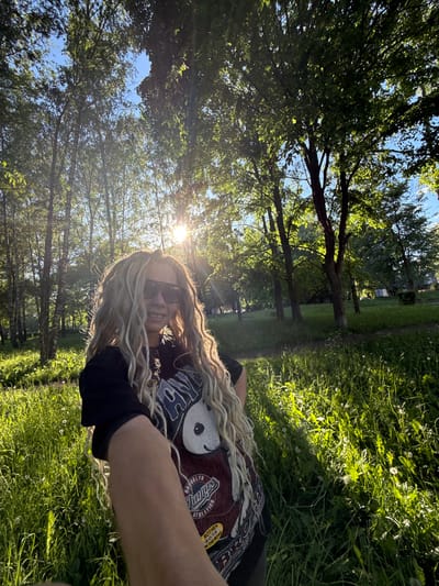 The image is a vertical full-frame selfie taken outdoors in a park or green space within the city of Rusanovo, Russia. The central subject is a person, appearing to be female, smiling broadly and looking directly at the camera. She wears dark sunglasses, obscuring her eyes, and has long, prominent light blonde and multi-toned dreadlocks or braids that frame her face and extend over her chest and shoulders. She is wearing a dark short-sleeved t-shirt with a maroon section featuring a graphic of a black-and-white figure and text including "BROOKLYN CHAMPS ATHLETICS" and a yellow patch with "ROTHER B.O.T.R ON THE ROAD". Her right arm is visible in the lower right foreground, extending towards the camera.
The environment is lush and green, characteristic of a sunny day. Strong sunlight originates from the upper right background, creating a distinct sunburst and lens flare, backlighting the subject and brightly illuminating the surrounding foliage. The foreground is covered in tall, green grass, interspersed with white dandelion seed heads.
The background features a dense canopy of deciduous trees with vibrant green leaves, through which patches of clear blue sky are visible. Some trees exhibit lighter bark consistent with birch species on the left side, while others have darker trunks. In the far background, particularly on the right, less dense foliage suggests a more open area or a path, potentially with faint indications of distant structures.
