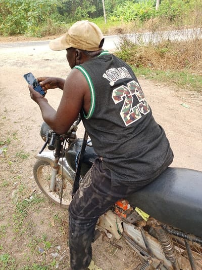 An African man wearing a tan baseball cap and a black Michael Jordan tank top sits on a motorcycle, looking at his phone. The motorcycle is old and shows signs of wear and tear. The man is wearing black pants. The background shows a dirt road with vegetation on either side. A paved road is visible in the distance.