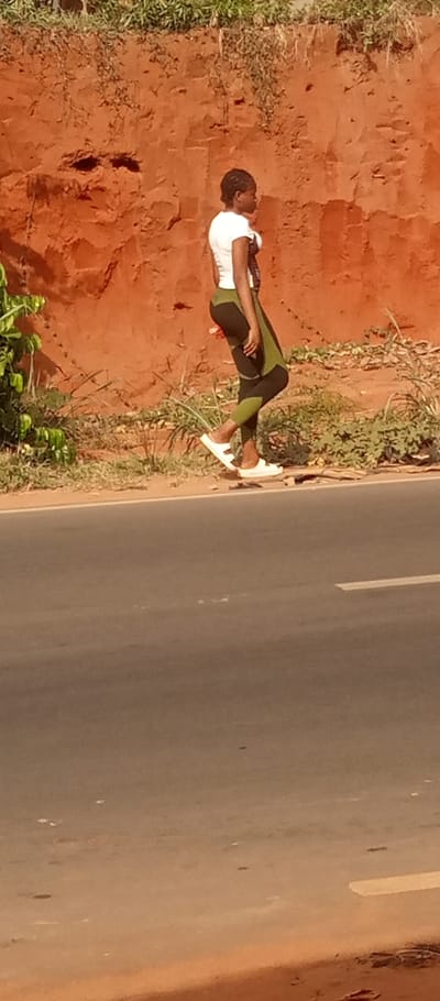 An outdoor scene in Ogidi, Nigeria, depicts a lone individual walking leftward along the edge of a paved road. The individual is a female, seen in profile, wearing a white short-sleeved shirt, olive green and black patterned leggings, and white flat sandals. Her hair is styled in dark braids. To her left, partially obscured, is green foliage. Behind her, extending across the upper two-thirds of the image, is a distinct reddish-brown earth embankment, uneven in texture, with sparse green and dry vegetation growing on its surface and at its base. The paved road surface occupies the lower third of the image, featuring a faint white broken line marking. The lighting suggests daylight conditions.