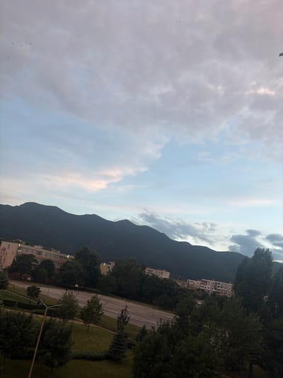The image depicts a scenic view of Vratsa, Bulgaria, likely taken from a high vantage point. The foreground is dominated by a vast expanse of sky filled with dramatic, wispy clouds, hinting at a partly cloudy or overcast day. The clouds are illuminated by soft, diffused light, suggesting it might be late afternoon or early evening.

In the mid-ground, a dark, imposing mountain range stretches across the frame, its slopes covered in lush, green trees. Nestled at the base of the mountain are several residential buildings, characterized by their white facades and numerous windows, indicative of an urban or suburban settlement. The buildings appear to be apartment complexes, suggesting a populated area.

A road with visible lane markings winds its way through the scene, bordered by a well-maintained green lawn and more trees. The road surface appears somewhat wet, possibly from recent rain. The overall atmosphere is serene and tranquil, with a sense of natural beauty juxtaposed against the built environment. There is no visible text within the image.