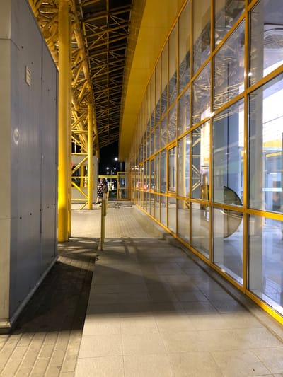 A person stands on a paved walkway next to a building with large yellow-framed windows. The walkway is bordered by a grey wall on the left and the glass-fronted building on the right. Yellow support beams and a yellow-painted roof structure are visible above the walkway. The ground is paved with rectangular tiles and square tiles. Shadows from the yellow supports and roof structure stretch across the walkway. The scene appears to be an exterior walkway, possibly at a transportation hub or public building, illuminated by artificial lights. The location is identified as Starovoitove, Ukraine.