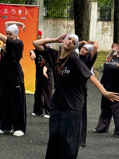 In an outdoor setting with a blurred background of trees and a building, a group of people are engaged in what appears to be a dance or performance. The subjects are dressed in black clothing, with several individuals wearing white headscarves. One person in the foreground wears a white headscarf and a black long-sleeved shirt, with their face partially visible and looking towards the camera. Another person in the foreground is also wearing a black outfit and a white headscarf, with their hands in a stylized pose. Behind them, other individuals in similar black attire and head coverings are also positioned in dynamic poses.
One individual in the background, wearing a red beanie, looks out from behind an orange banner that has several logos and text in Russian. Visible text on the banner includes "КОЛЕСО" and other unreadable characters. The overall atmosphere suggests a daytime event, possibly a street performance or a dance rehearsal, taking place on a paved surface. The people appear focused on their activity, with some showing concentration and others a more neutral expression. The scene is captured from a slightly elevated perspective, looking down at the subjects.