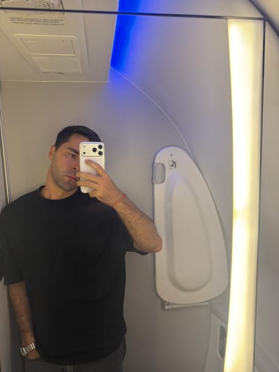 A man wearing a black t-shirt, grey pants, and a watch is taking a selfie with a white smartphone inside an airplane cabin. Overhead storage is visible with a white door and a light fixture. The environment appears to be an airplane cabin with overhead storage and interior lighting. No indications suggest this is in Tzafria, Israel.