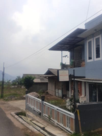 The image depicts a static, outdoor scene in Morrumbala, Mozambique, under an overcast sky. In the foreground, the edge of a paved or dirt road is partially visible on the left, alongside a concrete drainage ditch containing light-colored pipes or debris. A white and brown/tan fence with dark posts borders a property on the right. Dominating the right side of the frame is a two-story light blue building featuring white-framed windows on the upper floor. This upper level has a balcony protected by a dark-colored overhead canopy supported by thin posts; multiple hanging planters are visible on the balcony. On the ground floor of this building, a section with a dark wall and glass elements is present. A white rectangular sign with unreadable text hangs from a black pole positioned near the fence line in front of the building. To the left and slightly behind the main building, a smaller, single-story structure with a dark, sloped roof and lighter walls is visible, appearing to have a metal or wooden door. Further left, a utility pole with overhead wires extends across the frame. Sparse vegetation, including small trees and bushes, occupies the ground between the structures and extends towards faint, distant mountains or hills on the horizon. No individuals are present in the scene.