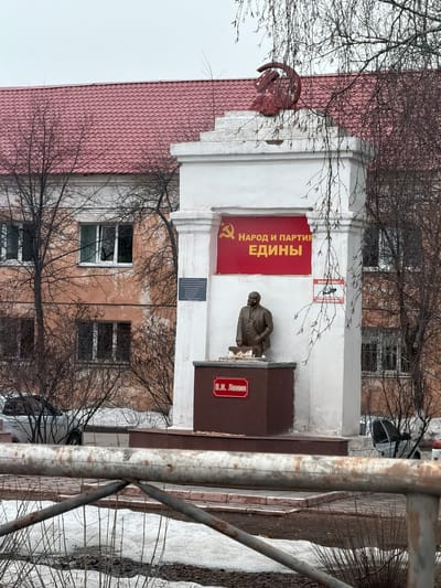 The image depicts a monument to V.I. Lenin located in Beloyarsky, Russia, observed under an overcast sky. The central feature is a white, multi-tiered concrete monument topped with a prominent red hammer and sickle emblem. Directly below the emblem, a red banner displays the yellow Russian text "НАРОД И ПАРТИЯ ЕДИНЫ" (The People and Party are United), accompanied by a smaller hammer and sickle symbol. A bronze bust of V.I. Lenin, portrayed from the waist up in a suit with his left hand extended, is mounted on a dark brown pedestal at the base of the white structure. A red plaque on this pedestal reads "В.И. Ленин". A rectangular dark plaque is affixed to the left side of the white monument, and a smaller sign with green elements is positioned on its right side. In the background, a multi-story building with a faded peach-colored facade, multiple white-framed windows, and a red corrugated metal roof is visible. Bare deciduous trees with sparse branches partially obscure the background building and frame the monument. The ground exhibits patches of melting snow interspersed with damp, dark earth. A light-colored vehicle is partially visible parked behind the monument on the left. A rusty metal barrier is present in the immediate foreground, horizontally traversing the lower portion of the frame.