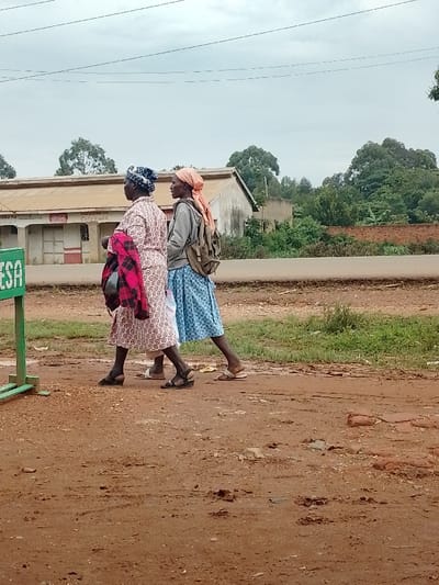 Two women, one wearing a pink and white dress and a blue head covering, the other wearing a blue and white dress and an orange head covering, walk along a dirt road in Kivaywa, Kenya. The road is muddy, and there is a green sign in the foreground. The location is near the junction of the Kivaywa-Nambale road and the Kivaywa-Kisumu road. Behind the women, there are buildings with a red-tile roof, trees, and a brick wall. There are power lines overhead.