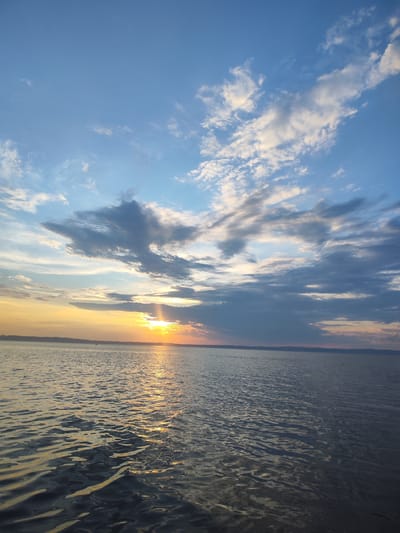 This image depicts a tranquil body of water, likely a lake or a large river, under a sky filled with scattered clouds. The sun is low on the horizon, casting a warm golden glow across the water and illuminating the undersides of the clouds with hues of orange and pink. The water's surface is gently rippled, reflecting the colors of the sky and the sunlight. In the distance, a faint, dark landmass is visible, suggesting a shoreline with some vegetation. The overall atmosphere is serene and peaceful, characteristic of a sunset or sunrise scene. No specific landmarks or details identifiable to Galevo, Russia are present in this generic landscape image.