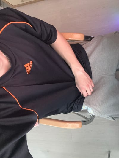 The image shows a person sitting in a chair. The person is wearing a black t-shirt with an orange Adidas logo and orange trim, and grey sweatpants. The background appears to be a room with light-colored walls and floor. The image is taken from a high angle, looking down at the person. There is no information in the image that can be used to determine the location in the city of Barnaul, Russia.