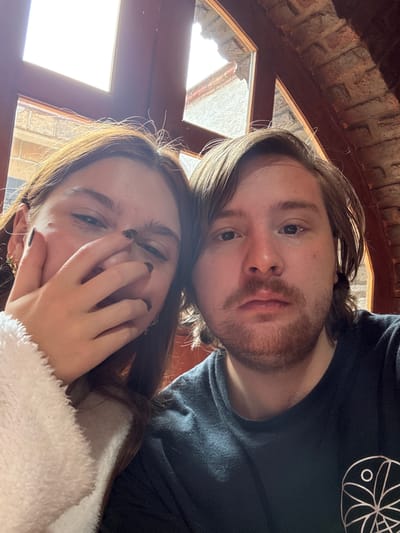The image is a selfie of a man and a woman, possibly taken in Cañada de Madero, Mexico, though no specific landmarks are visible. The man is positioned slightly above and behind the woman. He has light brown hair and a beard. He is wearing a dark t-shirt with a circular design on the upper right. The woman is in the foreground, with light brown hair and is wearing a white, fuzzy jacket. She has one hand partially covering her face. The background consists of an interior space with a window showing a bright outdoor scene and the ceiling is decorated with what appears to be brick.