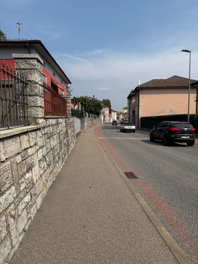 The image depicts a street scene in Balerna, Switzerland. On the left, a low stone wall borders a grey asphalt sidewalk. The wall is topped with a black metal fence and a red-painted railing. Behind the wall is a building with a red facade and grey upper sections, topped with a grey roof and a television antenna. On the right side of the street, a peach-colored building with a tiled roof stands adjacent to the road. A streetlamp is positioned in front of this building. The road itself is asphalt with red painted markings. Several vehicles are visible: a black Kia Sportage with a Swiss license plate (TI 99586) is in the foreground on the right, a white Peugeot is driving away in the middle distance, and another black car is further down the road. Palm trees and other greenery are visible along the right side of the street. The sky is blue with scattered white clouds. The scene appears to be set during the daytime.