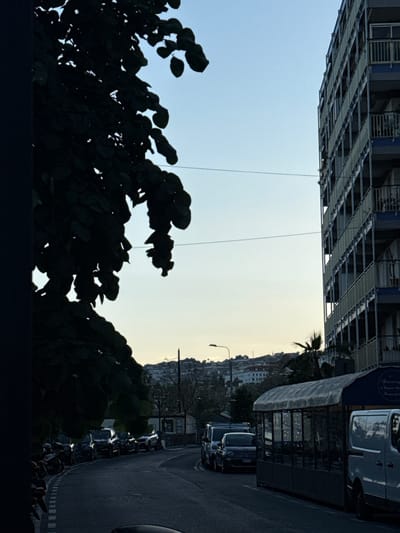 This is an outdoor scene in Sanremo, Italy, with a high-rise building under construction on the left. The sky transitions from blue to a pale yellow-white, indicating either sunrise or sunset. Dark foliage dominates the lower portion of the image, silhouetted against the sky. On the right side of the image, the view shifts to a street with parked vehicles and a storefront with a blue awning. Further into the distance, there are buildings on a hillside. A street lamp is visible near the street.