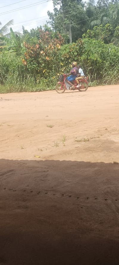 The image depicts a sandy road in Amauzu, Nigeria. A woman wearing a headwrap and blue clothes is driving a red motorbike with a young person in a white shirt riding pillion. The road is lined with vegetation. The foreground is shadowed.