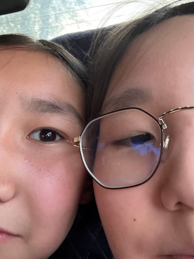 The image is a close-up, eye-level shot of two young Asian females, likely sisters, in a seated position, possibly in a vehicle. Their faces are positioned side-by-side, with their heads touching. The female on the left is looking directly at the camera, her left eye visible. She has natural eyebrows and fair skin with some freckles. The female on the right is wearing glasses and is looking slightly away from the camera, with her right eye partially closed. The glasses have thin, gold-colored arms and octagonal lenses. The background is blurred and shows what appears to be the rear window of a vehicle with a hint of trees or foliage outside. The lighting is natural, coming from above and slightly in front of the subjects. There are no identifiable landmarks or clues within the image that would indicate the location is Bishkek City, Kyrgyzstan.
