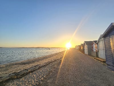 The image depicts a coastal scene within Poole, United Kingdom, likely along Poole Harbour, at either sunrise or sunset. A bright, low-lying sun is positioned slightly left of the image's vertical center in the mid-ground, casting a prominent lens flare diagonally across the upper-right sky. The sky is clear, transitioning from light blue at the top to yellow and orange hues near the horizon. The left half of the frame shows a calm body of water with gentle ripples, reflecting the sky's colors. Distant landmasses or structures, including a small vessel and several wooden posts, are visible on the far horizon across the water. A narrow strip of sandy and pebbly beach, with darker, wet sections and some debris, borders the water from the lower-left to the mid-ground. Adjacent to the beach on the right, a dark, granular paved or gravel walkway extends into the distance, parallel to the shoreline. A continuous row of beach huts lines the right side of this walkway. The foremost hut, located in the lower-right foreground, is dark blue or grey with light-colored double doors. Subsequent huts visible along the row display varying colors, including white, grey, and one with vertical red and white stripes. No individuals or active subjects are present within the frame.