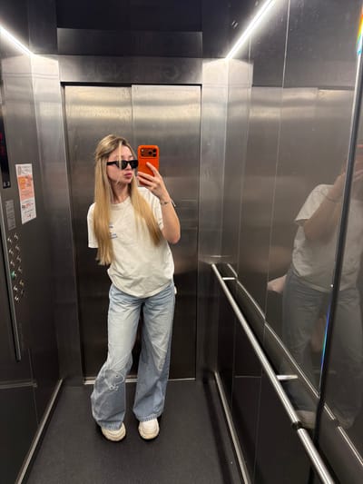 A young female stands centered inside a metallic elevator, capturing a selfie using an orange-cased smartphone held in her right hand. She has long, straight blonde hair, wears black rectangular sunglasses, a light-colored crew-neck t-shirt, and light blue wide-leg jeans with white outlined graphics of the Playboy bunny visible on the upper left leg. Her footwear consists of white sneakers. Her lips are pursed for the photograph.
The elevator interior features brushed metallic walls. The right wall acts as a reflective surface, showing a partial, inverted reflection of the subject. Overhead, a continuous LED strip light illuminates the space. The left wall contains a control panel with multiple circular buttons and a digital display that partially shows "PISO" (Portuguese for "floor") and numbers. Below this panel, the brand name "SCHINDLER" is embossed in black. The floor is dark and appears textured. A metallic handrail is affixed to the right wall. Inverted text, "CLÍNICOR", is visible on the upper section of the back wall, reflected in the right wall. This detail places the location within the Clínicor - Centro Médico de Reabilitação, situated at R. de Sá da Bandeira 786, 4000-444 Porto, Portugal.