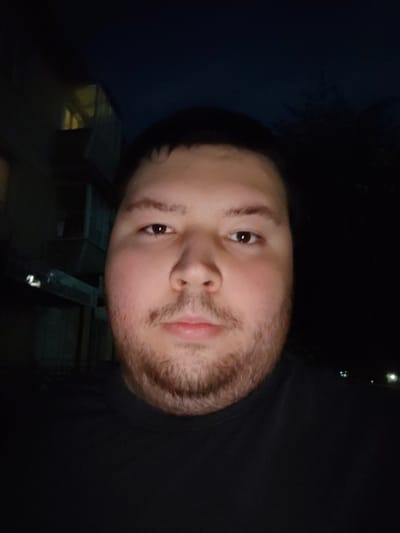 A selfie taken at night shows a close-up of a man's face with short, dark hair and light brown eyes. He has a fair complexion and a light beard. The man is wearing a black t-shirt. In the background, there is a dark building with illuminated windows and some trees. The image appears to have been taken outdoors in the city of Vladimir, Russia, given the presence of architectural elements suggestive of a residential building and the dark, unlit environment typical of a Russian city at night.