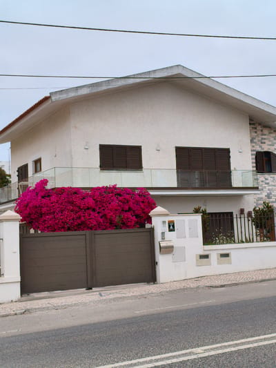 The image shows a modern house in Nazaré, Portugal. The house is white with a tiled roof and has dark brown shutters on the windows. In front of the house is a large hedge of vibrant pink bougainvillea flowers, which partially obscures a dark brown metal gate and fence. The street in front of the property is paved and appears to be in good condition. The sky is overcast, suggesting a cloudy day. There are no people or vehicles visible in the image, and the scene appears to be peaceful and well-maintained.