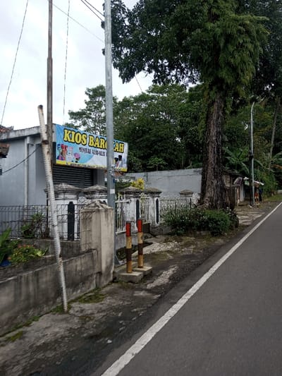 The image depicts a roadside scene, identified by a banner as "JL. WARUNG CISE, KAB. BANDUNG," placing the location in Bandung, Indonesia. The sky is overcast.
In the left foreground, a low concrete retaining wall with sparse vegetation leads to a decorative, white and black concrete and metal fence. Behind this fence is a light-grey, single-story building with a closed corrugated metal roll-down door. A blue banner prominently displayed on the building reads "KIOS BAROKAH" in yellow letters, with "MENJUAL ALAT-ALAT..." (Sells tools/equipment...) and the aforementioned address below.
Multiple utility poles are present; a rusty metal pole stands on the far left, and a grey metal pole is centered in the midground, both supporting overhead lines. To the right of the building and fence, a large tree with a textured trunk and dense green foliage dominates the midground. Behind the fence and tree line, a grey concrete wall is visible, backed by lush green trees and vegetation.
The foreground features a grey asphalt road with a solid white line marking its edge. A dirt and muddy strip runs parallel to the road, containing two short, concrete-based bollards painted with alternating red/white and brown/yellow bands. Further down the road on the right, additional informal structures are visible, and two indistinct human figures are present in the distance. A street light pole is also visible further down the road on the right.