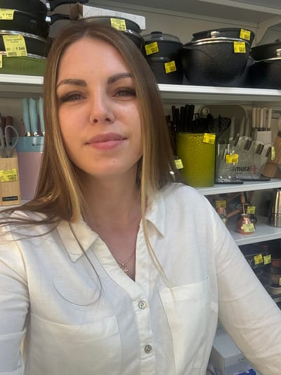 The image shows a woman with long, highlighted brown hair, wearing a white linen shirt with button closures. She is in what appears to be a cookware and kitchenware aisle of a retail store. Behind her is a shelf displaying a variety of items including black pots, knife blocks (one labeled "samura JAPAN"), and metal kitchen utensils. Price tags are visible on various items on the shelves. Text on a price tag indicates that the location is in Klin, Russia.