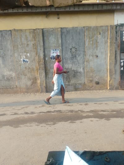 A young woman in a pink top and light blue denim skirt walks along a paved road. She's wearing sandals and holding a mobile phone. Behind her is a tall concrete wall with a faded yellow top. The wall has a few posters or signs on it and some decorative elements. The road surface appears worn, with patches and variations in texture. The photo appears to be taken during the daytime with natural lighting.
