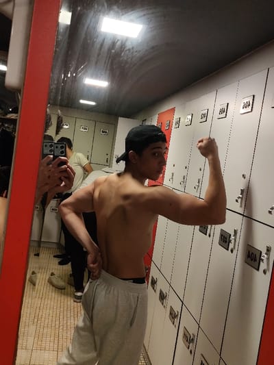 A young man is captured posing in what appears to be a locker room or changing facility, likely within Rukn ad-Din Municipality, Syria. The main subject is shirtless, wearing light grey sweatpants and a black baseball cap worn backward. He stands with his back partially to the camera, looking over his right shoulder towards a mirror, flexing his left bicep. His back and shoulder muscles are visible.
The setting features a prominent row of white lockers with silver handles and black numerical plaques on the right side of the image. Some lockers are accented with red panels. Visible locker numbers include "80A", "804", and "807". A large mirror, framed in red on the left, reflects the room and a second individual. This person, wearing a light-colored t-shirt, is visible in the mirror's reflection, holding a smartphone with multiple camera lenses, seemingly taking the photograph.
The ceiling above the lockers has rectangular fluorescent lights, which are also reflected in the mirror. The floor is tiled with a light-colored pattern. A pair of light-colored sneakers is visible on the floor in the reflection. The overall scene suggests a moment of displaying physical fitness, possibly after a workout.