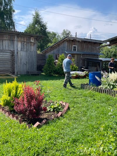 The image depicts a backyard scene in the village of Kumyonki, Kirov Oblast, Russia. The environment is rural, characterized by a wooden house, a garden with flowers and shrubs, and a grassy lawn. A man in camouflage pants and a blue t-shirt is walking towards the right side of the frame, appearing to be talking on his phone. A woman in a dark floral top stands in the background on the right, also facing towards the right. In the middle ground, a bright blue barrel stands near a small wooden fence. The garden features red and yellow shrubs, along with some white flowers. The overall setting suggests a casual outdoor gathering or activity in a residential area.