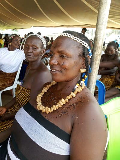 The image shows three older women sitting under a tent. The woman in the foreground is wearing a striped shawl and a necklace made of peanuts. She also has a headband made of white shells. Her hair is braided, and there are blue beads in the braid. Her face and shoulders are covered in small black seeds. The woman next to her is also wearing a peanut necklace and shell headband and a yellow patterned cloth. A third woman in the background is wearing a white shirt and similar yellow cloth. The women are sitting on plastic chairs. The location is likely in Takau, Nigeria, given the context provided. The tent is made of a light-colored fabric.