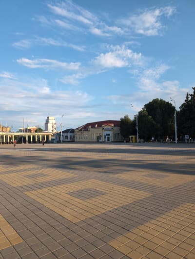 This image captures an outdoor scene in Tikhoretsk, Russia, specifically a wide, paved plaza in front of several buildings. The plaza's paving stones are laid in a contrasting pattern of yellow and gray squares, with long shadows cast across it, suggesting it is late afternoon or early morning.

In the background, a row of light-colored buildings with red-tiled roofs stands. One building prominently displays signage with Cyrillic text that appears to be for a bank or financial institution, reading "Кубань Кредит." To the left of these buildings, a long, covered walkway with white columns and yellow roofing is visible, leading towards a taller, modern-looking structure with a white tower. The sky above is a bright blue, dotted with scattered white, wispy clouds.

Several individuals are present in the plaza. One person is riding a bicycle on the left side of the image, while others are walking in smaller groups in the middle ground, near the buildings and walkway. Lush green trees are interspersed throughout the background, adding a natural element to the urban setting. The overall impression is of a public space in a Russian town, with a mix of architectural styles and everyday activity.