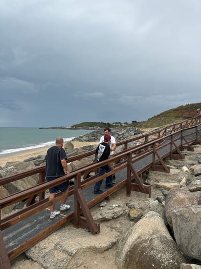 Three individuals are on a wooden ramp and walkway overlooking a beach and the sea. The man in the foreground, wearing a black t-shirt and blue patterned shorts, is walking away from the camera. To his left, two other individuals are facing each other and talking on the walkway. The person on the left has red hair, wears a black t-shirt with a white graphic, and dark pants. The person on the right wears a white t-shirt and dark pants. The walkway is constructed with brown wooden railings and a dark grey surface. The beach to the left is sandy with some rocks, and the sea is a greenish-blue with white waves. The sky is overcast with dark clouds. In the background, there is a slight incline covered with grass and brush, with some buildings and a harbor visible in the distance. The location is the ramp leading to the beach at Courtown, Ireland.