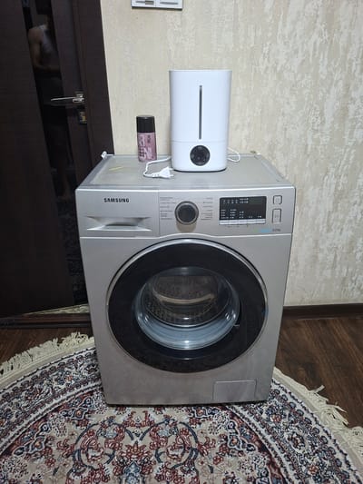 The image shows a silver Samsung washing machine placed on a circular rug. On top of the washing machine are a white humidifier and a dark pink spray can. The washing machine has a digital display and control panel on its front. The background consists of a beige textured wall and a dark brown door with a silver handle. A person's upper body is partially visible in the reflection of the door. The floor is made of wooden planks. The scene appears to be indoors, possibly in a residential setting in Kokand City, Uzbekistan.