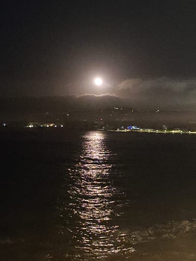 The image presents a tranquil night scene featuring a bright moon over a body of water, likely the Mediterranean Sea off Dénia, Spain. A prominent, luminous full or near-full moon is positioned in the upper center of the dark sky, partially obscured by soft, wispy clouds that diffuse its light.
The moonlight creates a striking, shimmering pathway across the water's surface, forming a glistening column of light that extends from the distant horizon towards the foreground. The water appears to be rippled, enhancing the reflective effect.
In the mid-ground, a distant coastline is visible, defined by numerous artificial lights that indicate a town or city. These lights are scattered across the land, with some concentrated along the immediate shoreline and others twinkling higher up on the hills behind. The blend of natural moonlight and distant urban illumination creates a serene yet captivating atmosphere.
The overall setting is dark and atmospheric, with no people or specific activities depicted. The scene evokes a sense of peace and the quiet majesty of a moonlit night over the sea. No text is visible in the media.