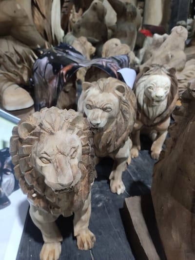 The image depicts a collection of wooden lion carvings. Three lion figures are positioned prominently in the foreground. The carvings are detailed, showcasing the lions' manes, facial features, and paws. The wood appears to be light in color and roughly textured. The background contains more wood carvings and unfinished wooden pieces. The scene appears to be inside a shop or workshop, possibly in Ubud, Indonesia, based on the style of woodcarving.