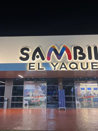 This is a night-time, eye-level, outdoor, medium shot of the Sambil El Yaque shopping mall in Pampatar, Venezuela. The Sambil logo, with its colorful "M", is prominently displayed above the entrance. Below the logo, the words "EL YAQUE" are illuminated. The mall features large glass doors and windows, reflecting the lights from inside and the surrounding environment. The ground in front of the mall appears wet, suggesting recent rain, and is a reddish-brown color. A silver trash receptacle is visible to the left of the entrance. Inside the mall, through the glass, various displays and store fronts are visible, with some signage including "SE VIVE EL" and "AMIGO". One large display features a white car and text that reads "Vive la pasión del fútbol en el mejor Centro comercial de la ciudad". A purple informational stand with white text is positioned to the left of the entrance.