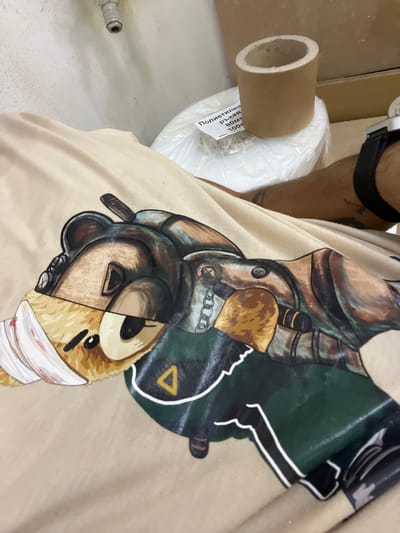 The image shows a close-up of a beige t-shirt with a graphic print of a character that appears to be a stylized bear wearing armor or a costume. The character has a bandaged left cheek, and its body is depicted in metallic and green tones, with some chain detailing and a yellow triangular emblem. The t-shirt is laid out on a flat surface, possibly a table or bed. In the background, out of focus, there are rolls of material, one wrapped in plastic and another of cardboard. A person's arm, with visible tattoos and a black watch, is also present in the background, resting on the t-shirt. The environment appears to be indoors, with a textured wall visible at the top left. There is no discernible indication of the city of Sevlievo, Bulgaria, in this image.
