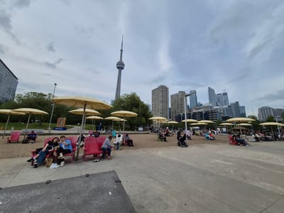 This is a wide-angle, eye-level, full shot of a beach area in Toronto, Canada, with the CN Tower visible in the background. The beach is covered in sand, with rows of red Adirondack chairs and yellow umbrellas providing seating and shade for beachgoers. In the foreground, a paved walkway runs along the edge of the beach. Several people are scattered throughout the scene, some sitting in chairs, others walking or pushing strollers. A couple in the left foreground is taking a selfie, while a man and child are in the middle ground. The background features a dense collection of modern skyscrapers, indicating an urban setting. The sky is overcast with scattered clouds.