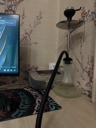An interior scene in Kurovskoye, Russia, depicts a workstation with a hookah on a light-colored wooden desk. A silver-stemmed hookah with a black bowl and a clear glass base, containing a translucent light green liquid and white vapor, occupies the right side of the desk. A black hose extends from the hookah's base, curving towards the viewer. To the left, a dark-framed monitor displays a video with Russian text: "Тьерри" and "a Footlit dit Footlit (в титрах: Thiérrée)", along with a timestamp of "-00:40:05" and video playback controls. Below the monitor, a dark mousepad with an abstract calligraphic pattern in teal, white, and pink tones is visible, with a black gaming mouse resting on it. Behind the mousepad, a silver-colored portable CD player or mini-stereo system is present, connected via an auxiliary cable. A white multi-port USB charger, featuring three visible USB ports, is plugged into an electrical outlet above and behind the audio device. The wall in the background is covered with light-toned wallpaper patterned with branches, leaves, flowers, and birds in muted colors. No individuals are present in the image.