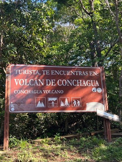 The image shows a sign for "VOLCÁN DE CONCHAGUA" in Amapalita, El Salvador. The sign, which is predominantly red, features white text and several white icons. The text "TURISTA, TE ENCUENTRAS EN:" is at the top, followed by "VOLCÁN DE CONCHAGUA" in larger letters. Below that, "CONCHAGUA VOLCANO" is written in smaller text. The icons depict a mountain with a rope, a person at a viewpoint, coniferous trees, and two hikers. There are also two stickers on the sign: one circular with "RIDERS" and "SURF" around an image, and another circular sticker with "EL SALVADOR" and a sun symbol. To the right of the main sign, a smaller wooden directional sign with white text pointing to the right reads "RUMBO AL ESPÍRITU DE LA MONTAÑA." The sign is situated outdoors, surrounded by lush green trees and foliage, suggesting a natural, possibly mountainous, environment. The ground in front of the sign is covered with dry leaves, dirt, and some green grass. The lighting indicates it is daytime, with sunlight filtering through the trees, casting shadows on the sign.