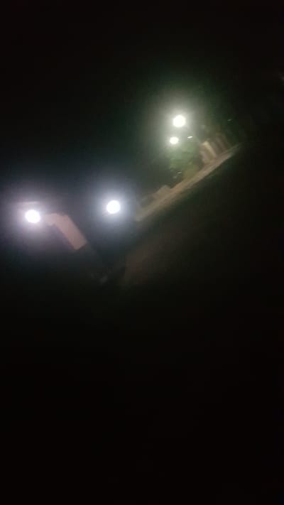 A nighttime scene in Wukari, Nigeria, is dimly lit by artificial lights. Two bright lights, possibly from a vehicle or streetlights, illuminate the left side of the image, casting a glow on what appears to be a building structure behind them. Further in the background, another set of two lights can be seen, also suggesting artificial illumination. The overall environment is dark, typical of a street or outdoor area at night. The scene is devoid of any visible people or animals, focusing solely on the interplay of light and shadow on the urban landscape.