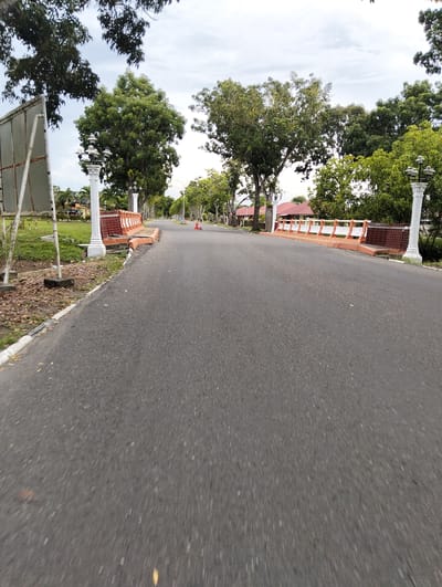 The image depicts an empty, paved road in Tambontunong, Indonesia, lined with decorative orange and white barriers, streetlights on ornate white pillars, and lush green trees. On the left side of the road, a white sign with a metal frame stands on a patch of grass. In the background, houses with red roofs are visible, interspersed with more trees. The sky is overcast. The road appears to be a quiet street or a section of a larger park or residential area.