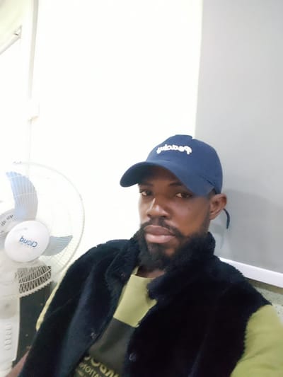 The image shows a close-up, eye-level, indoor shot of a Black man wearing a blue baseball cap and a black furry vest over a light green t-shirt. He has a beard and is looking towards the camera. To the left of the man, a white electric fan is visible. The background consists of a plain white wall and a grey section on the right. The overall setting appears to be an indoor space in Ikeja, Nigeria, possibly a home or office.