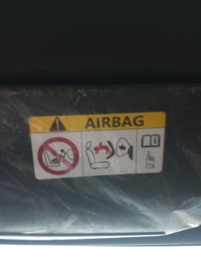 The image shows a close-up view of an airbag warning sticker on a car's sun visor. The sticker is yellow and white with black text and symbols. The text "AIRBAG" is clearly visible in the center of the sticker. To the left of the text is a warning triangle symbol with an exclamation mark. Below the text, there are three icons. The first icon shows a car seat with a circle and a diagonal line through it, indicating no objects or children on the seat. The second icon depicts a figure being ejected from a car seat, illustrating the force of an airbag deployment. The third icon shows an open book with an "i" inside, suggesting to consult the owner's manual. The sun visor appears to be covered in a protective plastic film. The background is dark and out of focus. There is no indication of the city of Pallawarukka, Indonesia, in this image.