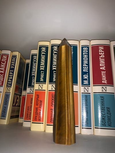 This media file shows a stack of books on a white shelf. The books have various titles in Russian, including "War of the Worlds," "The Old Man and the Sea," and "The Divine Comedy." The spine of one book, "The Divine Comedy," is partially visible at the top of the stack. The titles are printed in Cyrillic letters, and some have illustrations or logos on the spines. A golden object, possibly a pen or a bookmark, is placed across some of the books. The overall scene suggests a collection of literary works, possibly in a home library or a bookstore. The lighting is even and appears to be artificial, indicating the photo was likely taken indoors. The stack of books is neatly arranged, with the spines facing outward.