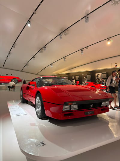 The image depicts the interior of the Museo Enzo Ferrari in Modena, Italy. A red Ferrari Testarossa is prominently displayed in the foreground, parked on a white platform. The car is angled to the right, with its front end facing the viewer. Behind it, another red classic Ferrari is visible, along with two people interacting with exhibits on a wall in the background. The ceiling features a track lighting system illuminating the space. The overall environment is clean and modern, typical of a museum exhibition.