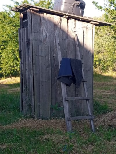 The image shows a rustic wooden outhouse with a black water tank on its roof, set amidst a grassy and overgrown rural landscape. A wooden ladder is propped against the outhouse, with a blue garment and a dark-colored fuzzy garment draped over its rungs. The outhouse itself is constructed from weathered, vertical wooden planks, and its roof appears to be made of corrugated metal or similar material. The surrounding environment consists of green grass, dry straw or hay, and trees in the background. The lighting suggests it is daytime, with shadows indicating the sun is at an angle. There are no people visible in the image. The scene appears to be in a rural area, possibly a backyard or a secluded spot. The image does not provide specific information about the city of Luka, Ukraine, beyond the general impression of a rural setting.
