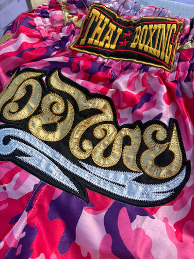 A close-up, overhead shot focuses on a pair of brightly colored camouflage Muay Thai shorts. The shorts feature a pink, purple, and lighter pink camouflage pattern. Prominently displayed on the front of the shorts are two embroidered patches. The upper patch, a black rectangle with a gold border and red trim, reads "THAI BOXING" in gold lettering, with two red stars between the words. Below this, a larger, more elaborate patch features gold, stylized lettering that appears to be Thai script, set against a black background. This script is outlined in gold, with a white, jagged design underneath. The fabric of the shorts is shiny and appears to be satin. The background is slightly out of focus, showing a neutral surface, possibly concrete or a similar material, suggesting an outdoor setting. The image does not show any people or specific landmarks. It is not possible to determine the location in Arugam Bay, Sri Lanka from the provided image.