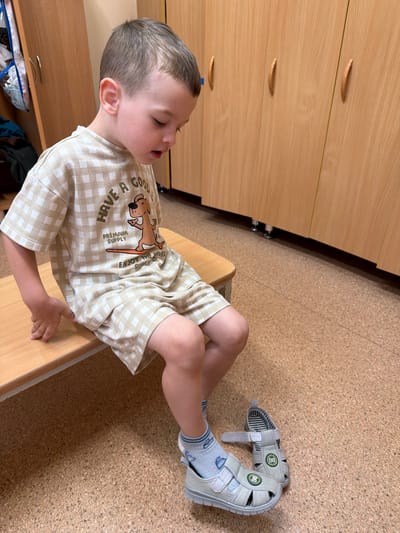 A young boy with short brown hair is sitting on a wooden bench, looking down at his feet. He is wearing a beige and white checkered t-shirt with a graphic of a dog on a surfboard and text that reads "HAVE A GOOD PREMIUM SUPPLY ENJOY THE WAVES". He is also wearing matching checkered shorts and light blue socks. One sandal is on his left foot, and the other is lying on the floor next to his right foot. The sandal on his foot is grey with green accents and the number "98" on it. The sandal on the floor is also grey with green accents and the number "86" on it. The boy is in what appears to be a locker room or changing area, with light brown wooden lockers visible in the background. The floor is covered in a textured brown material, possibly cork. The lighting is bright and even.