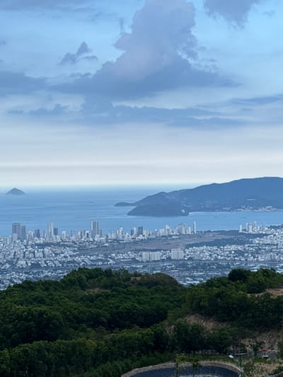 An aerial view shows the coastal city of Nha Trang, Vietnam, near Diên Khánh. The ocean and islands separate the city from overcast skies on the left. Dense green foliage lines the foreground on the right. Buildings and infrastructure populate the land between the water and the trees.